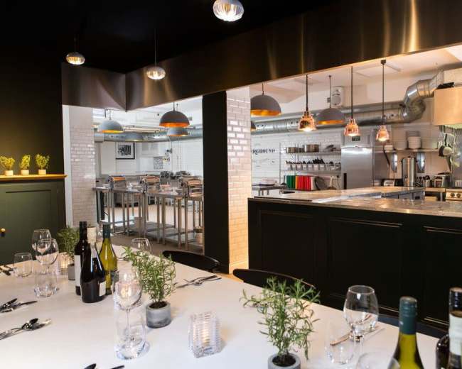 A modern kitchen with stainless steel appliances is visible behind a dining area set with a white tablecloth, wine bottles, and decorative herbs.