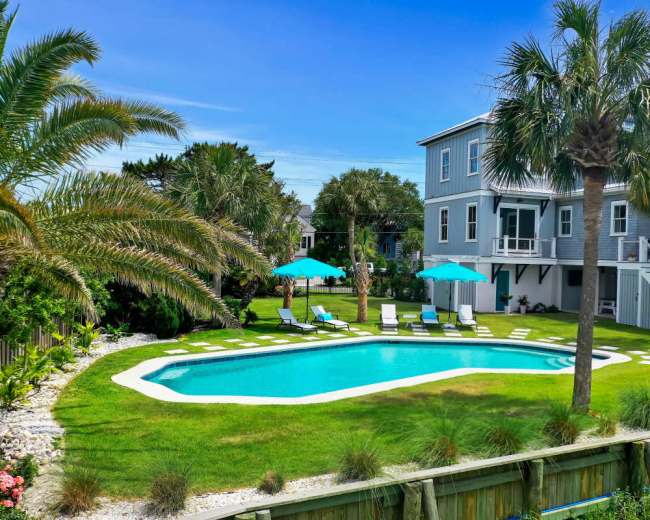 The image shows a private swimming pool surrounded by palm trees and lounge chairs, with a two-story house in the background.
