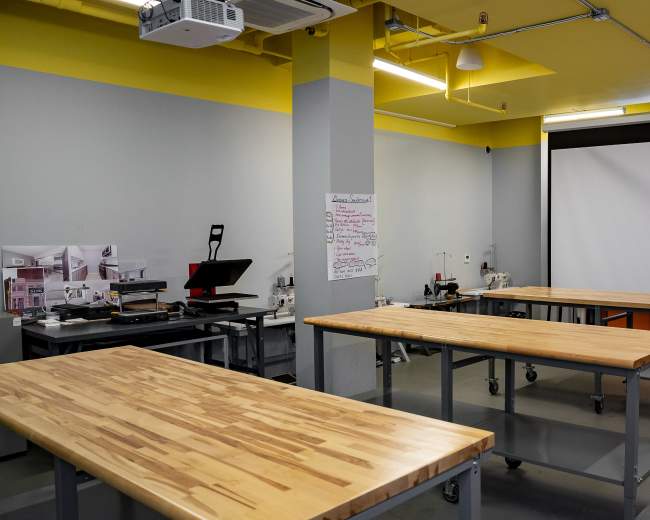 A spacious workshop is equipped with wooden tables, a projector screen, and various tools on counters and shelves.