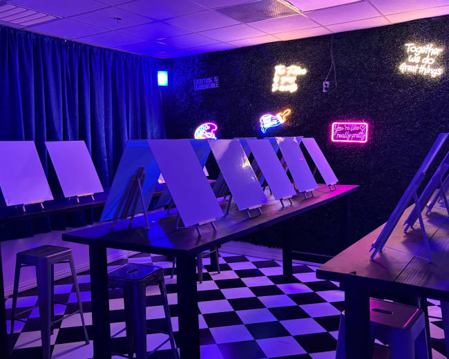 An art studio features several blank canvases on easels set against a vibrant, neon-lit wall.