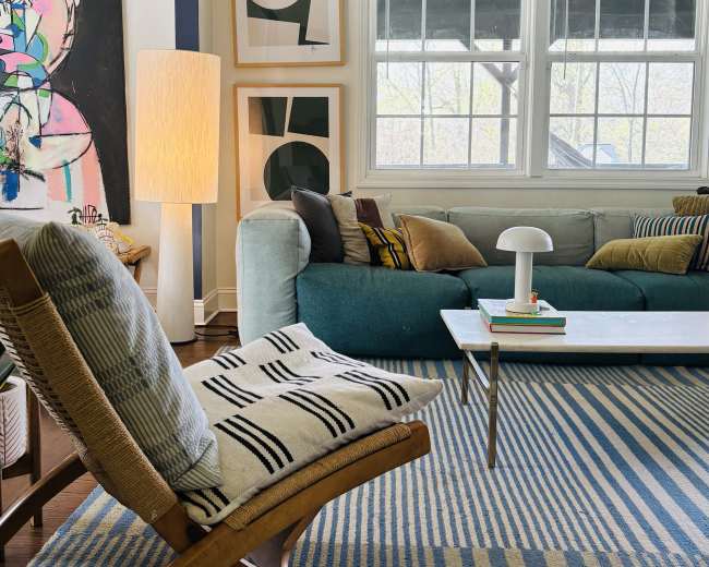 The image shows a cozy living room featuring a teal sofa, a patterned rug with blue stripes, a wooden chair, and abstract wall art.