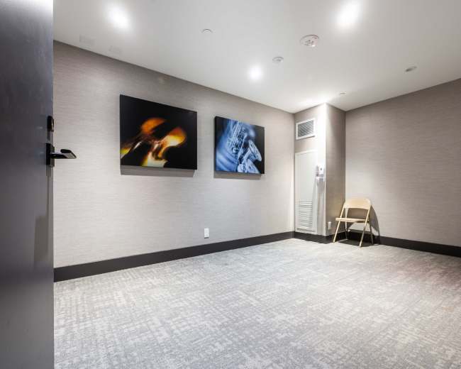 The image shows a small, empty room with two framed artworks on the wall and a single folding chair positioned against the wall.