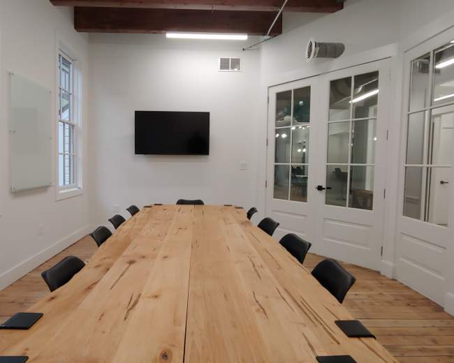 A long wooden table is set up in a brightly lit conference room, with chairs arranged around it and a large screen mounted on the wall.