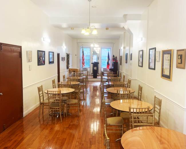 The image shows a well-lit dining hall with wooden floors, several round tables arranged with chairs, and framed documents and photos on the walls.