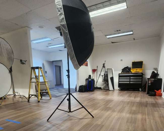 The image shows a spacious studio or room with wooden flooring, a large lighting umbrella on a stand, a ladder, and various equipment in the background.