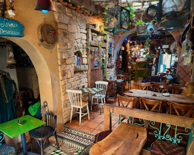 The image shows a colorful, rustic interior of a café or restaurant with wooden tables, ornate tiled floors, and various decorative items hanging from the walls.