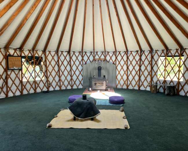 The interior of a yurt features a round, open space with wooden beams, a central wood stove, and circular seating arrangements on a green carpet.