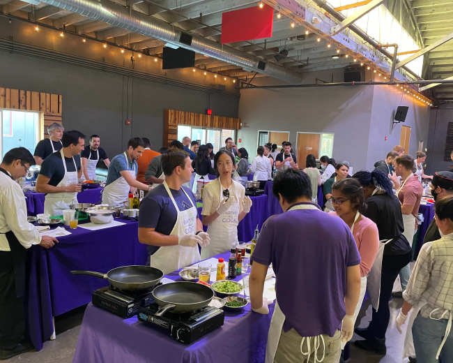 A large cooking event takes place in a spacious venue with participants gathered around tables, preparing various dishes.