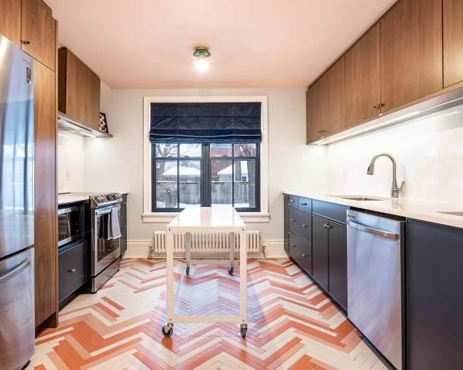 A modern kitchen features stainless steel appliances, dark cabinetry, and a chevron-patterned floor with a central table.