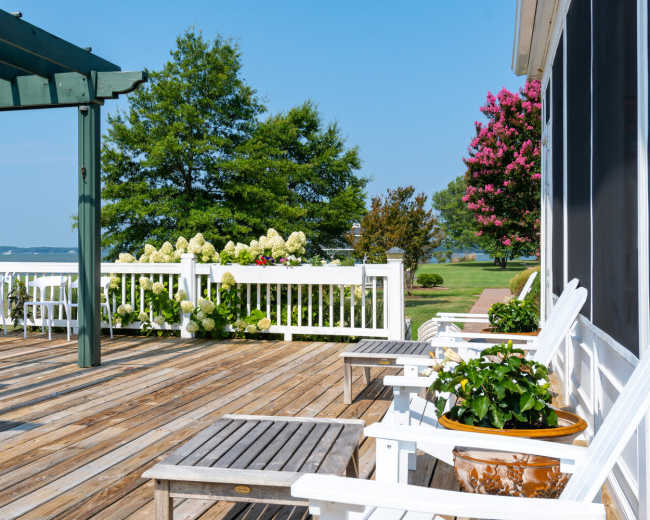 The image shows a wooden deck with white patio furniture and potted plants, overlooking a body of water surrounded by greenery.