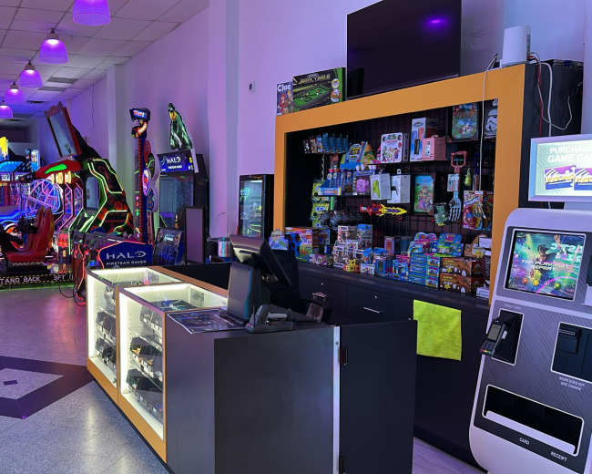 The image shows an arcade with various gaming machines on one side, a counter with merchandise and prizes, and vibrant lighting throughout the space.