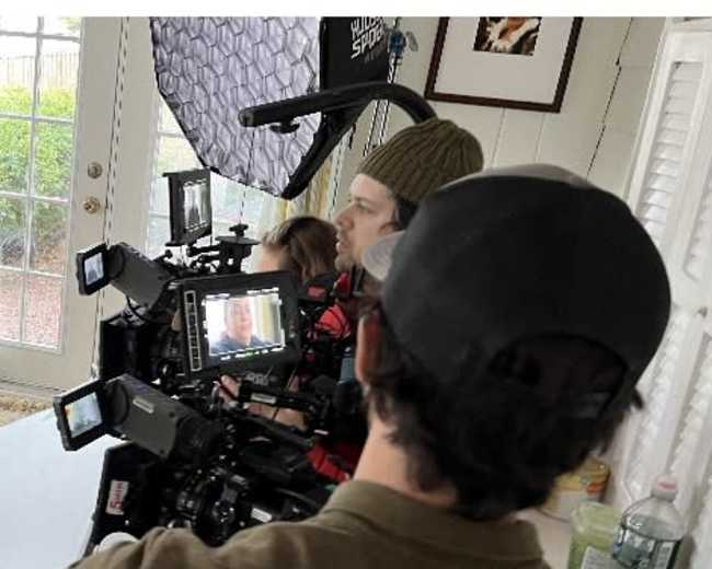 The image shows a film crew setting up multiple cameras and monitors in a room with natural light.