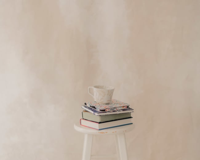 A stack of books with a decorative teacup sits atop a white stool against a light, textured background.