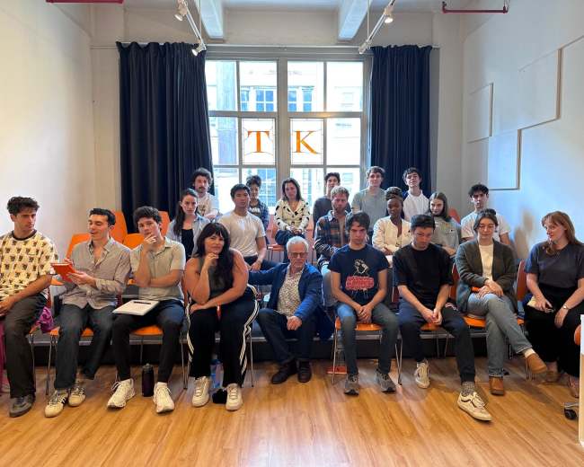 A group of twenty people, consisting of mostly young adults, is seated in a bright room with large windows and modern furnishings.