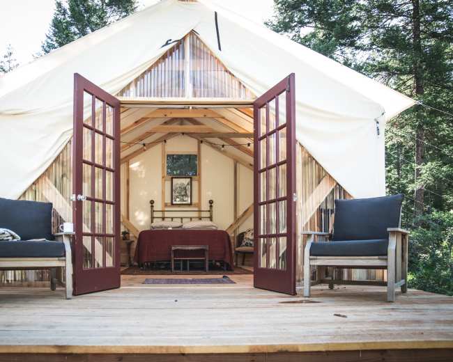 A canvas tent with wooden double doors opens to a rustic interior featuring a bed and two chairs on a wooden deck surrounded by trees.