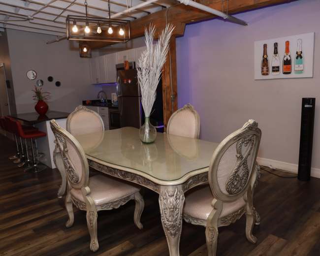 A glass table with ornate chairs is set in a dining area featuring a modern kitchen in the background and decorative wall art.