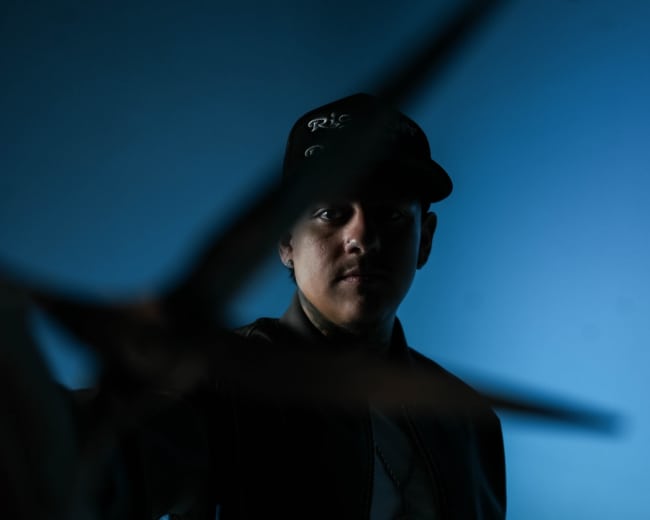 A person wearing a baseball cap and a dark jacket extends a pair of scissors toward the camera against a blue backdrop.