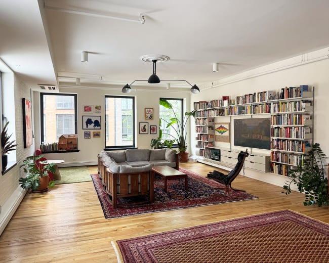 The image shows a spacious living room with a gray sectional sofa, wooden coffee table, and a bookshelf filled with books, surrounded by green plants and artwork on the walls.