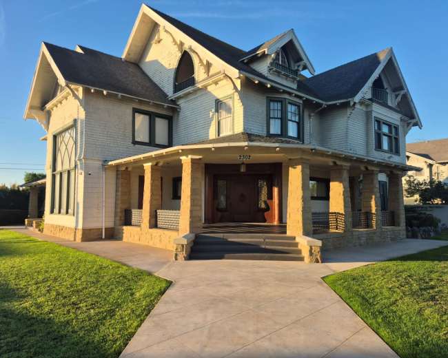 The image shows a large, two-story mansion with a white exterior, decorative elements, and a front porch supported by stone columns, set in a well-maintained yard.
