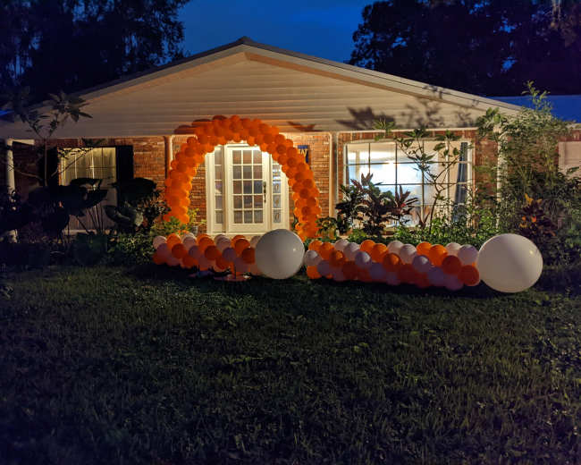 A house is decorated with an arch of orange and white balloons positioned in the front yard, illuminated in the evening light.