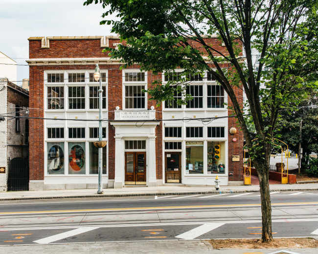 The image shows a two-story brick building with large front windows and a tree on the sidewalk, located across a street with painted crosswalks.