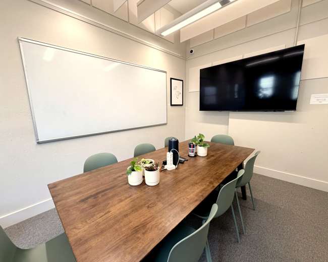 The image shows a conference room with a large wooden table surrounded by green chairs, a whiteboard on one wall, and a television screen mounted on another wall.