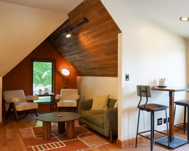 The image shows a cozy interior space featuring a small seating area with a sofa and chairs, a coffee table, and a dining setup near a window.