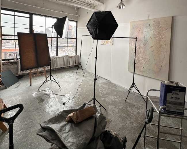 The image shows an empty art studio with two photography lights, a large window, and several art pieces displayed, including an abstract painting on the wall.