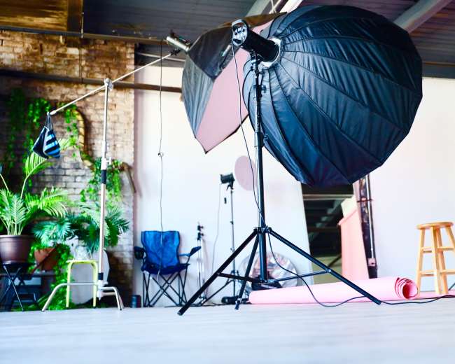 A photography studio with lighting equipment, a wooden stool, and a pink backdrop rolled on the floor.