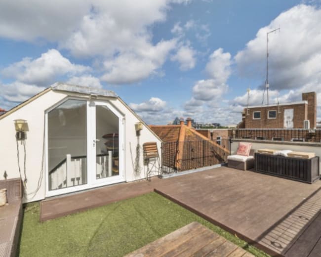 The image shows a rooftop terrace with seating areas, a wooden deck, and a view of surrounding buildings under a partly cloudy sky.