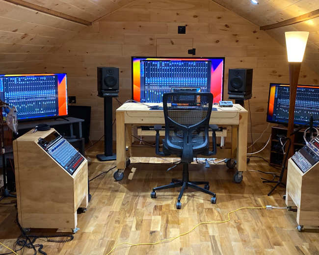 The image shows a home recording studio with three monitors, an audio mixing desk, and various audio equipment arranged around a central chair.