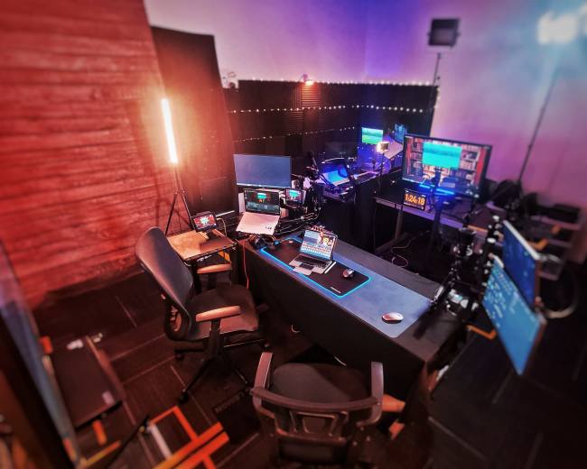 The image shows a small studio setup with multiple computers, monitors, and lighting equipment arranged around a central desk.