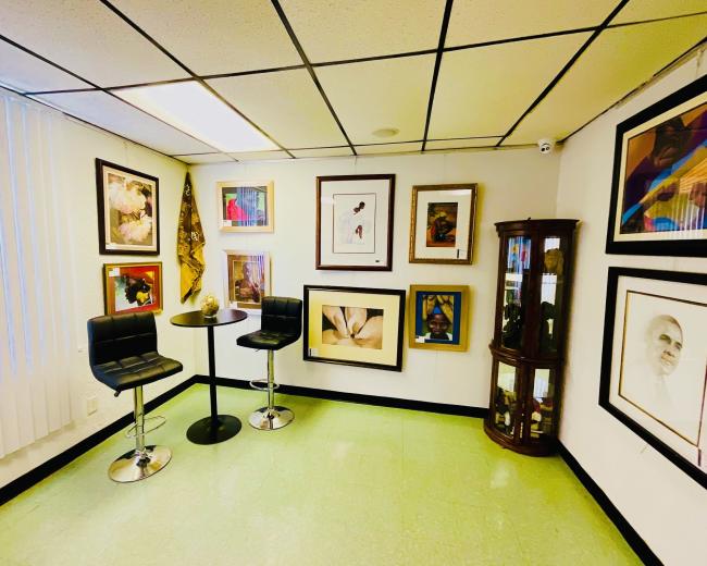 A small gallery room features a set of framed artworks on the walls, a round table with two black stools, and a display cabinet in one corner.