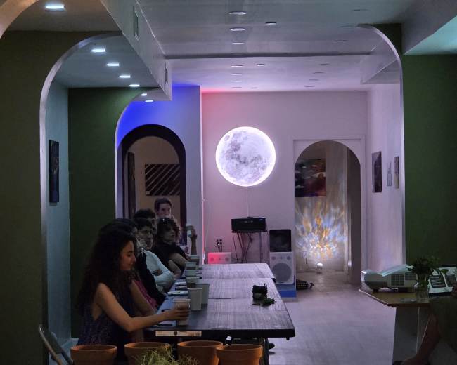 The image shows a modern indoor space with a long table and several people seated, illuminated by colorful lighting and featuring a large moon-shaped light on the wall.