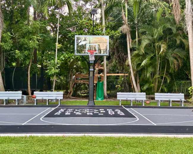 A basketball court surrounded by lush greenery features a hoop, a nearby slide, and benches for seating.