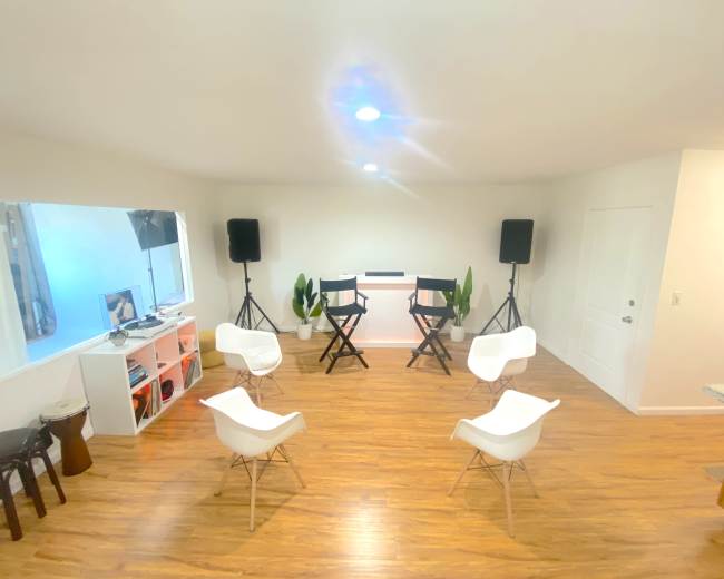 The image shows a spacious room with two director's chairs facing a small stage, surrounded by several white chairs and equipped with speakers and plants.