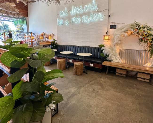 The interior features a cozy seating area with wooden benches, pendant lights, tropical plants, and a neon sign that reads, "eat desserts, be a monster."