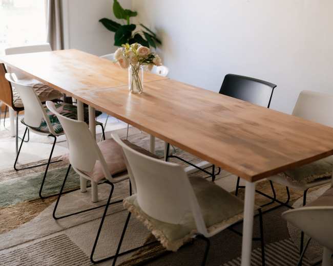 A long wooden table is set in a bright room surrounded by several white and black chairs, with a vase of flowers at the center and plants adorning the walls.