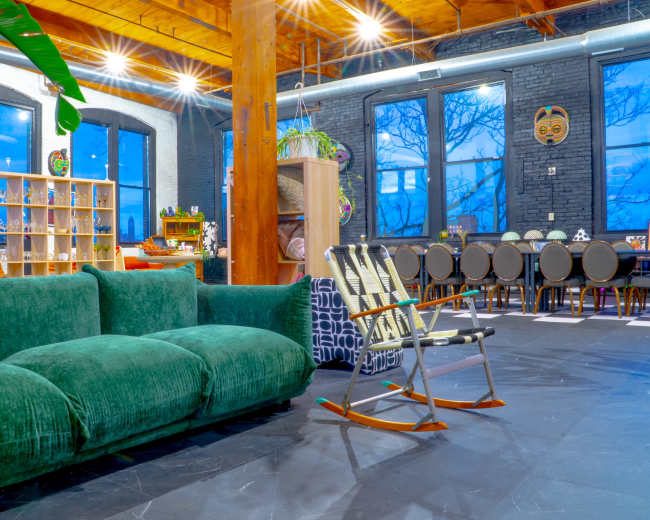 The image shows a spacious interior with a green velvet sofa, wooden beams, and a dining area in the background.