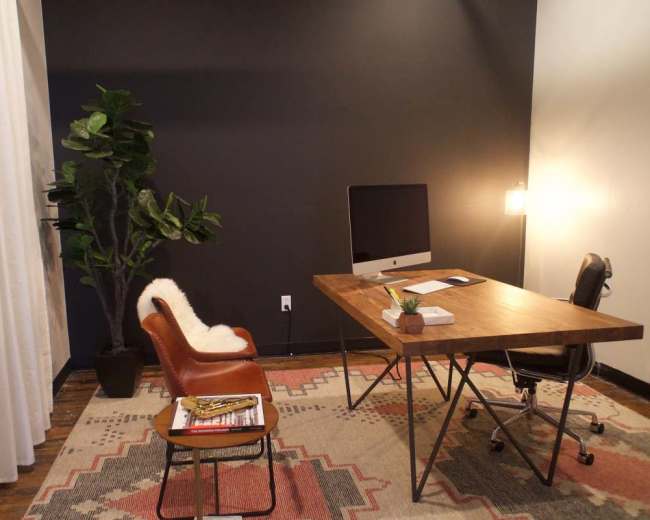 The image shows a modern office space featuring a wooden desk with an iMac, a brown leather chair, a potted plant, and a patterned rug on a wooden floor.