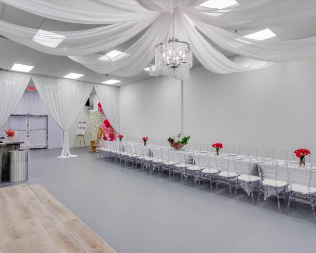 A spacious event venue with a long table set for dining, decorated with red flowers, beneath draped white fabric and a chandelier.