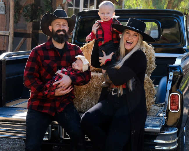 A family of four, dressed in matching plaid outfits, poses with two children in front of a pickup truck parked near hay bales.