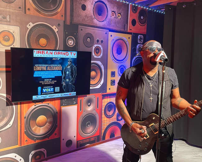 A musician performs with a guitar on stage in front of a large display featuring an event poster.