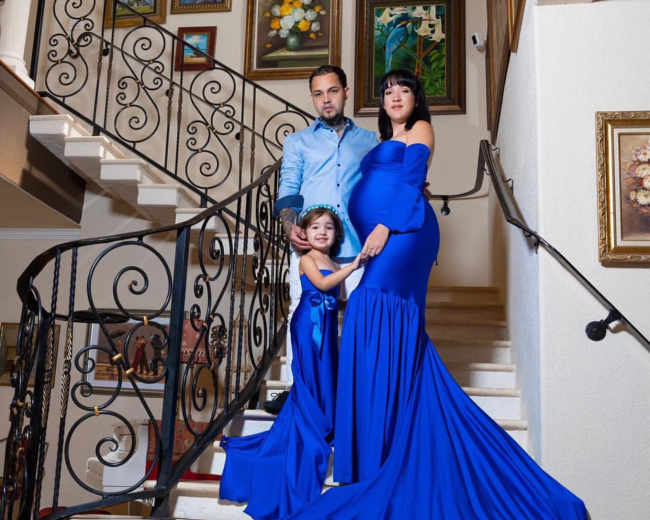 A man, woman, and young girl stand on a staircase, with the woman wearing a long blue dress that trails behind her.