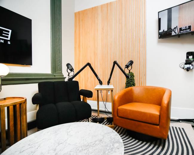 The image shows a cozy room featuring a black upholstered chair and an orange leather armchair, a circular marble table, and a wall-mounted television displaying a timer, against a backdrop of wooden paneling and a green-framed window.