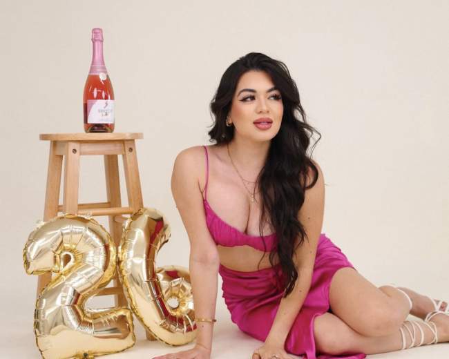 A woman in a pink dress poses beside a wooden stool holding a bottle of rosé and two large gold balloons displaying the number "20."