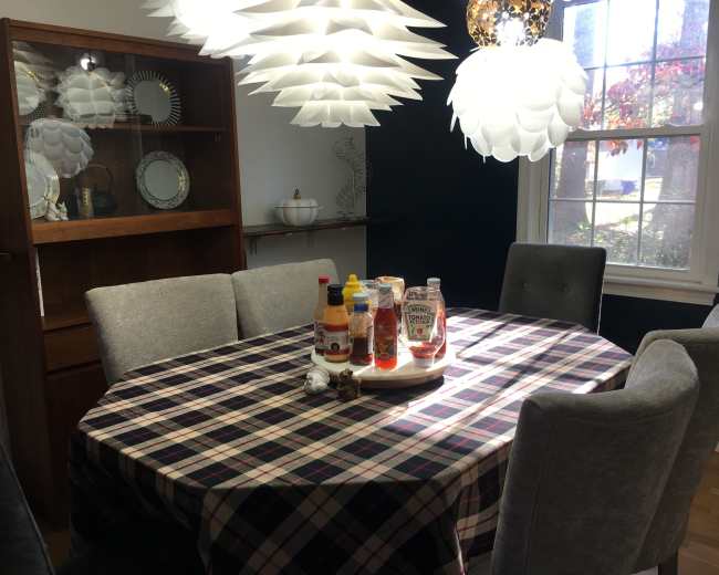 A round dining table with a plaid tablecloth is set with various condiments, surrounded by gray upholstered chairs, in a well-lit room featuring modern pendant lights and a wooden cabinet.
