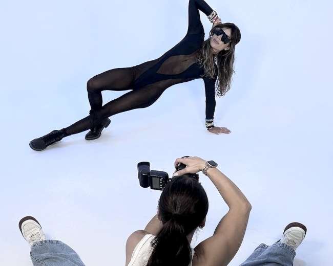 A model poses in a black bodysuit on a white backdrop while a photographer captures the shot from a seated position.