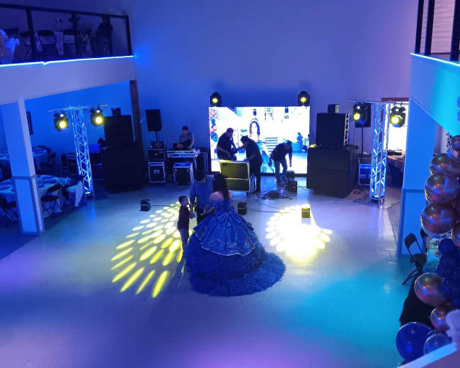 A brightly lit event space features a large screen, sound equipment, and a person in a vibrant blue dress performing on the floor.