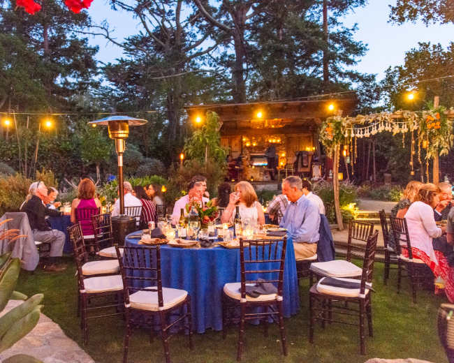 A garden dinner party is set up with tables and chairs, illuminated by string lights and surrounded by greenery.